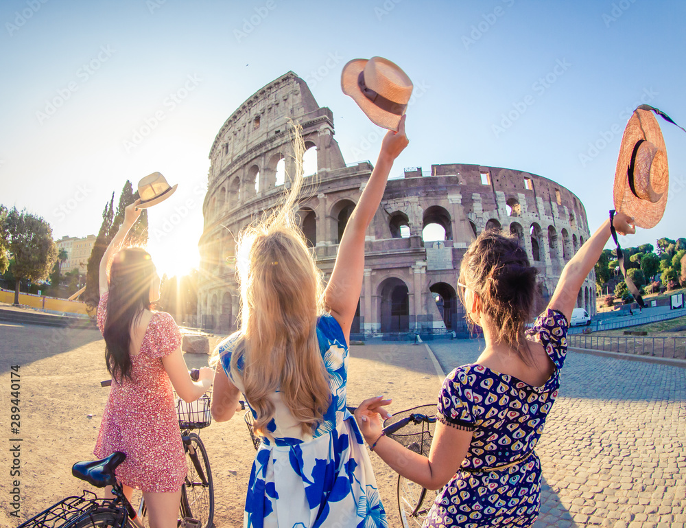 Three happy young women friends tourists with bikes waving hats at Colosseum in Rome, Italy at sunrise. - obrazy, fototapety, plakaty 