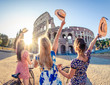 Three happy young women friends tourists with bikes waving hats at Colosseum in Rome, Italy at sunrise.