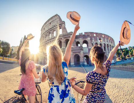 three happy young women friends tourists with bikes waving hats at colosseum in rome, italy at sunri