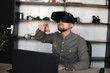 © Volodymyr - Young bearded man sitting against computer with virtual reality glasses trying to touch something or tap some thing in front of what he seeing. Modern technologies. The concept of future technology.