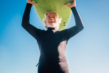 Happy Senior With Surftable On His Head Is Smiling And Laughing - Old And Mature Man Having Fun Surfing With A Black Wetsuits - Active Retired Adult Doing Activity Alone