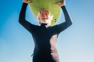 happy senior with surftable on his head is smiling and laughing - old and mature man having fun surfing with a black wetsuits - active retired adult doing activity alone