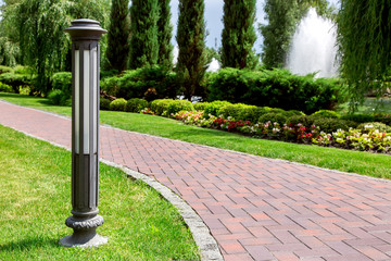 Wall Mural - ground decorative iron lantern in the park with footpath of red stone tiles and landscaping with flower beds and bushes with trees on a sunny summer day.