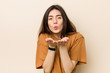 © Asier - Young brunette woman against a beige background folding lips and holding palms to send air kiss.