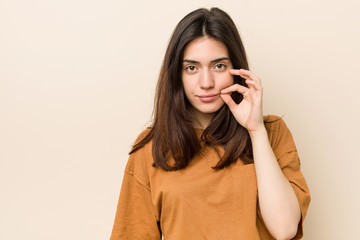 Wall Mural - Young brunette woman against a beige background with fingers on lips keeping a secret.