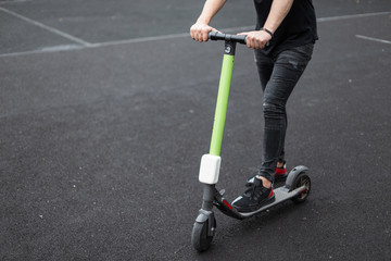 Poster - Fashionable young man in denim stylish clothes in sneakers holds hands on the steering wheel an electric scooter. Male legs standing on a modern scooter  on a basketball court. Close-up. Lifestyle.