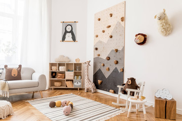 Stylish scandinavian interior design of childroom with gray sofa, modern climbing wall for kids, design furnitures, soft toys, teddy bear and cute children's accessories. Home decor. Template. 