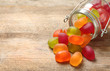 Glass jar and scattered jelly candies on wooden table, space for text