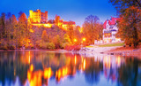 Fototapeta Nowy Jork - Germany, Bavaria. Autumnal scenic view of illuminated historic castle Schwangau, reflected in Alp See lake during twilights. Hohenschwangau castle circled by colorful fall trees. Popular landmark.