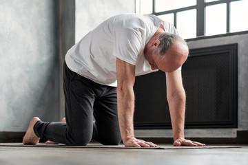 Senior bald yogi men practices yoga asana marjariasana or cat-cow pose at home