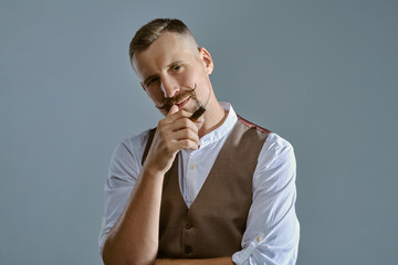 Wall Mural - Man with a stylish mustache, dressed in brown vest and white shirt is posing sitting on a table, enjoying cigar. Grey background, close-up.