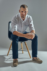 Wall Mural - Man with stylish mustache, in classic white shirt and blue trousers is sitting on chair, holding glass of whiskey. Grey background, close-up shot.