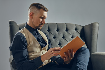 Wall Mural - Man with stylish mustache, dressed in black shirt and trousers, beige vest is sitting on dark sofa, reading a magazine. Grey background, close-up.