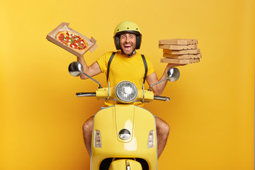 Wall Mural - Our most delicious pizza for you. Cheerful young man holds opened pizza box, drives yellow moped, being in hurry to transport fast food in time, wears protective helmet, has much duties at work