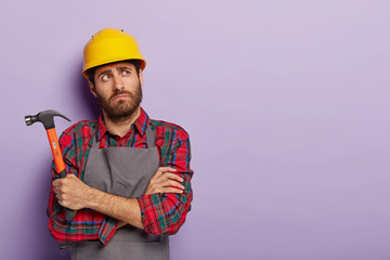 Photo of thoughtful tired mannual worker holds hammer, keeps arms crossed over chest, thinks about what to repair, wears yellow protective headgear, shirt with apron, copy space on purple background