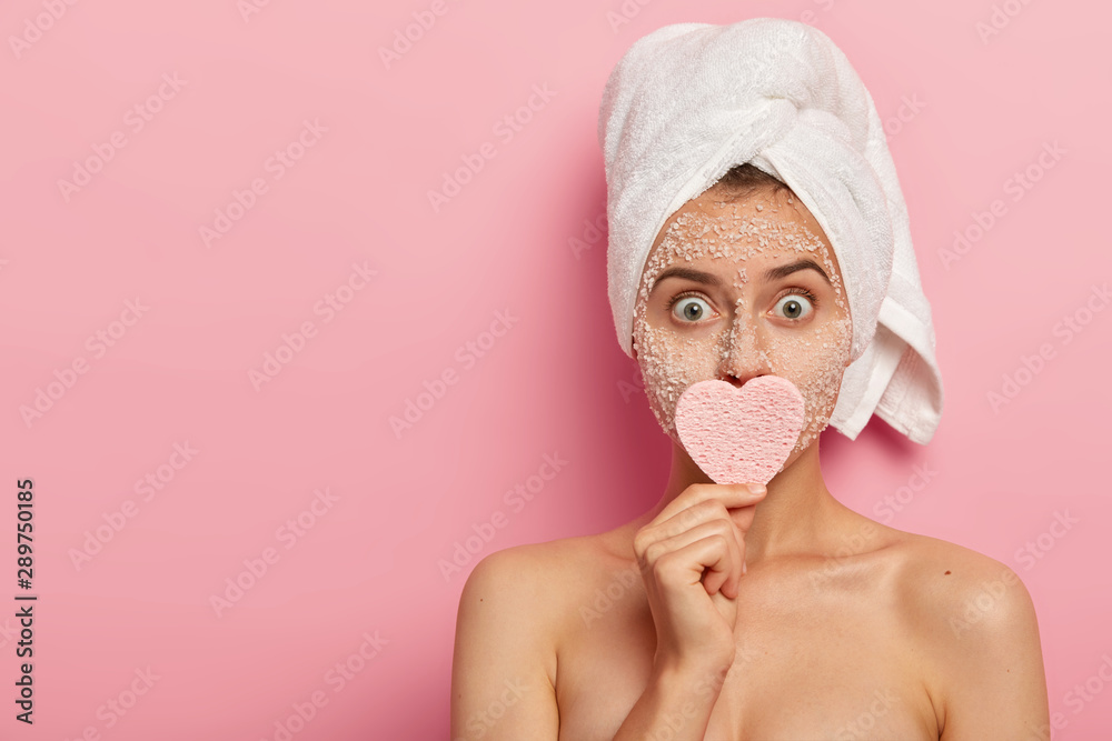 Studio shot of young woman has widely opened eyes, feels shocked, reduces dryness of skin with natural nourishing mask, keeps cosmetic sponge on mouth, busy with skincare routine, isolated on pink - obrazy, fototapety, plakaty 
