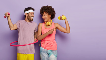 Horizontal shot of happy diverse couple have active sport training, raise dumbbells, rotate hula hoop, dressed in active wear, look joyfully at each other, listen music in headphones during workout