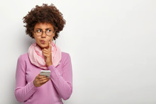 Pensive Dark Skinned Woman Looks Thoughtfully Aside, Holds Mobile Phone, Waits For Call, Purses Lips, Has Afro Haircut, Dressed In Violet Jumper With Scarf, Keeps Index Finger On Cheek, Poses Indoor