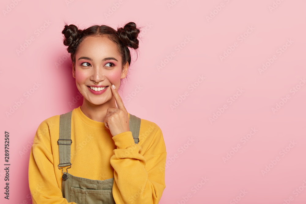 Pretty young woman with Asian appearance, wears yellow sweatshirt and overalls, has two hair buns, looks aside, models against pink background, blank space for promo. Emotions and beauty concept - obrazy, fototapety, plakaty 