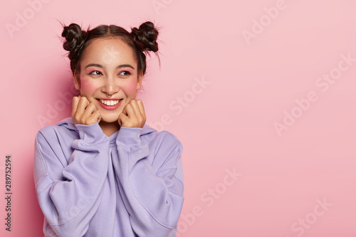 Studio shot of pretty smiling Asian woman with dark hair, two knots, wears purple loose jumper, stands against pink background, copy space area for your advertisement, expresses good emotions