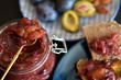 Plum chutney or jam in a glass jar with open lid and golden spoon on black tabletop. Slices of brown bread and plate with plums in blurred background. Selective focus.
