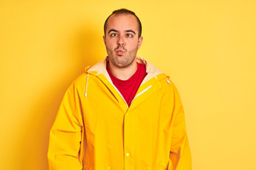 Sticker - Young man wearing rain coat standing over isolated yellow background making fish face with lips, crazy and comical gesture. Funny expression.