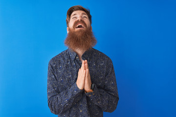 Canvas Print - Young redhead irish man wearing floral summer shirt standing over isolated blue background begging and praying with hands together with hope expression on face very emotional and worried