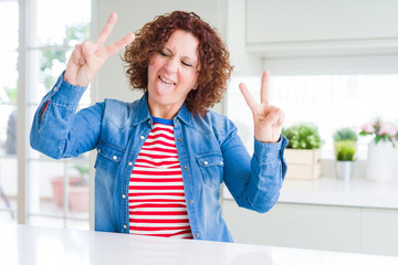 Poster - Middle age senior woman with curly hair wearing denim jacket at home smiling with tongue out showing fingers of both hands doing victory sign. Number two.