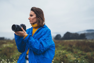 Wall Mural - photographer girl in blue raincoat hold in hands photo camera take photography foggy mountain, traveler shoot autumn nature, video click on camera technology, landscape vacation concept free space