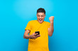 © luismolinero - Young man with yellow shirt over isolated blue background with phone in victory position