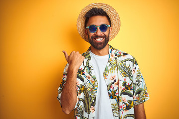 Wall Mural - Indian man on vacation wearing floral shirt hat sunglasses over isolated yellow background smiling with happy face looking and pointing to the side with thumb up.