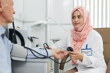 Portrait of young Arab woman working as doctor in medical clinic and measuring blood pressure of senior patient, copy space