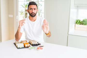 Poster - Handsome hispanic man eating asian sushi using chopsticks with open hand doing stop sign with serious and confident expression, defense gesture