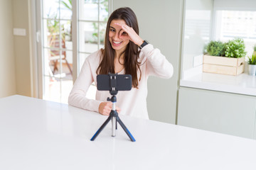 Poster - Beautiful young woman recording selfie video with smartphone webcam doing ok gesture with hand smiling, eye looking through fingers with happy face.