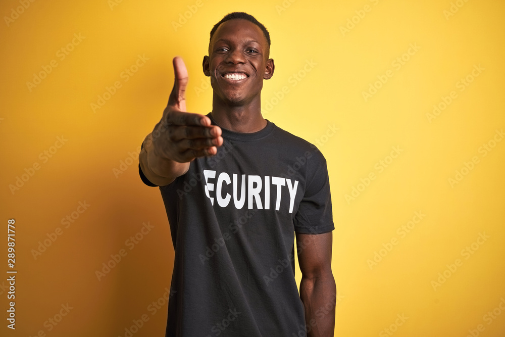 African american safeguard man wearing security uniform over isolated yellow background smiling friendly offering handshake as greeting and welcoming. Successful business. - obrazy, fototapety, plakaty 