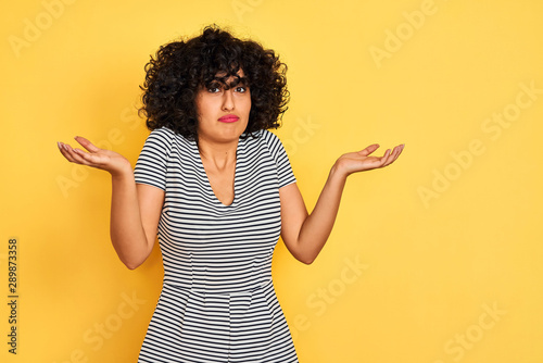 Fotografía Young arab woman with curly hair wearing striped dress over isolated yellow background clueless and confused expression with arms and hands raised