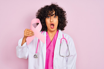 Sticker - Young arab doctor woman with curly hair holding cancer ribbon over isolated pink background scared in shock with a surprise face, afraid and excited with fear expression