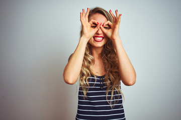 Wall Mural - Young beautiful woman wearing stripes t-shirt standing over white isolated background doing ok gesture like binoculars sticking tongue out, eyes looking through fingers. Crazy expression.