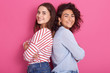 Profile of two attractive adult girls standing back to back, wearing casual clothing, looking smiling directly at camera, posing isolated over pink studio, females expressing happyness and joy.
