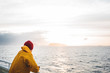 Hipster traveler wearing red hat and yellow raincoat looking away at cloudy mountain and sunset sea. Alone man traveling  at scandinavian authentic ocean landscape by ship