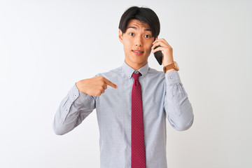 Wall Mural - Chinese businessman wearing tie talking on the smartphone over isolated white background with surprise face pointing finger to himself