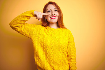 Sticker - Beautiful redhead woman wearing winter sweater standing over isolated yellow background Pointing with hand finger to face and nose, smiling cheerful. Beauty concept