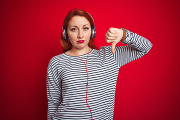 Wall Mural - Young redhead woman listening to music using headphones over red isolated background with angry face, negative sign showing dislike with thumbs down, rejection concept