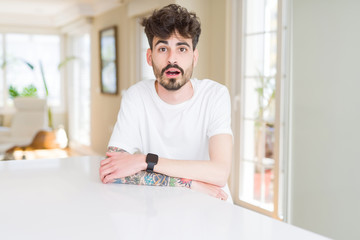 Poster - Young man wearing casual t-shirt sitting on white table afraid and shocked with surprise expression, fear and excited face.