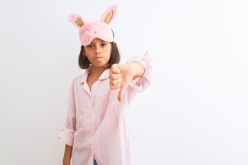 Wall Mural - Beautiful child girl wearing sleep mask and pajama standing over isolated white background looking unhappy and angry showing rejection and negative with thumbs down gesture. Bad expression.