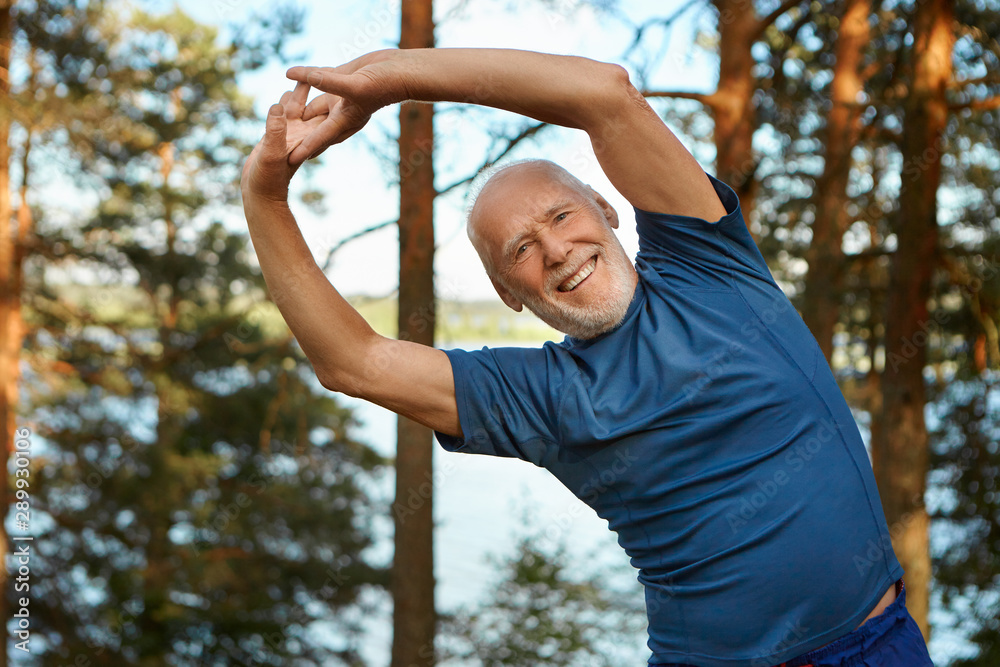 Outdoor shot of happy energetic senior retired man enjoying physical  training in park, doing side bends exercise, holding hands together,  looking at camera with broad smile, warming up body before run Stock
