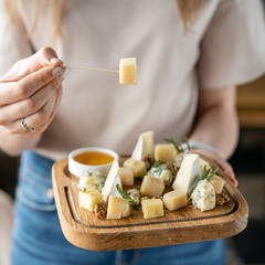 Girl holding piece of parmesan cheese on a skewer and wooden plate with cheese. Delicious cheese mix with walnuts, honey. Food for wine.