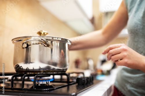 Cuadros en Lienzo Beautiful young woman housewife prepairing dinner, hold in hands big steel saucepan, standing it on gas-stove