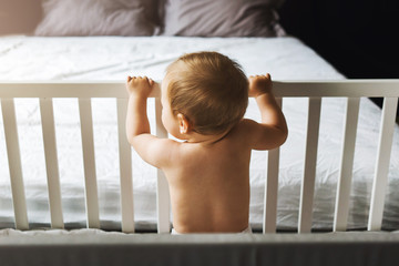 Rear view. A child alone is standing at home in a crib, holding on to its side and looking at an empty bed in the background.