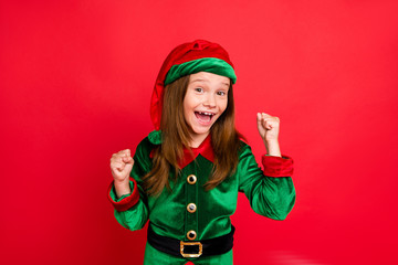 Poster - Portrait of shocked elf kid in green hat cap with long red head hairstyle raising her fists celbrate newyear party wearing fairy costume isolated over red background